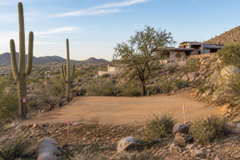 Staked homesite pad of clean decomposed granite with salvage-tagged saguaros at the pad edge and survey flags on a boulder-strewn slope